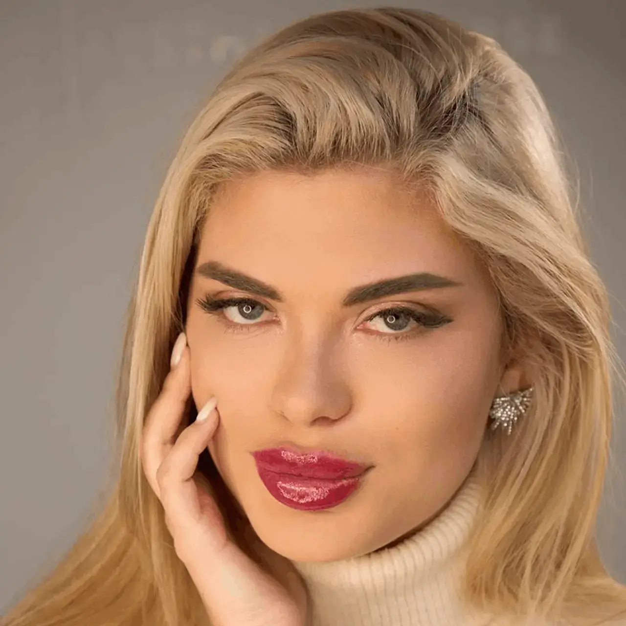 Studio portrait of a blonde woman with glossy red lips in a cream turtleneck, hand resting on her cheek