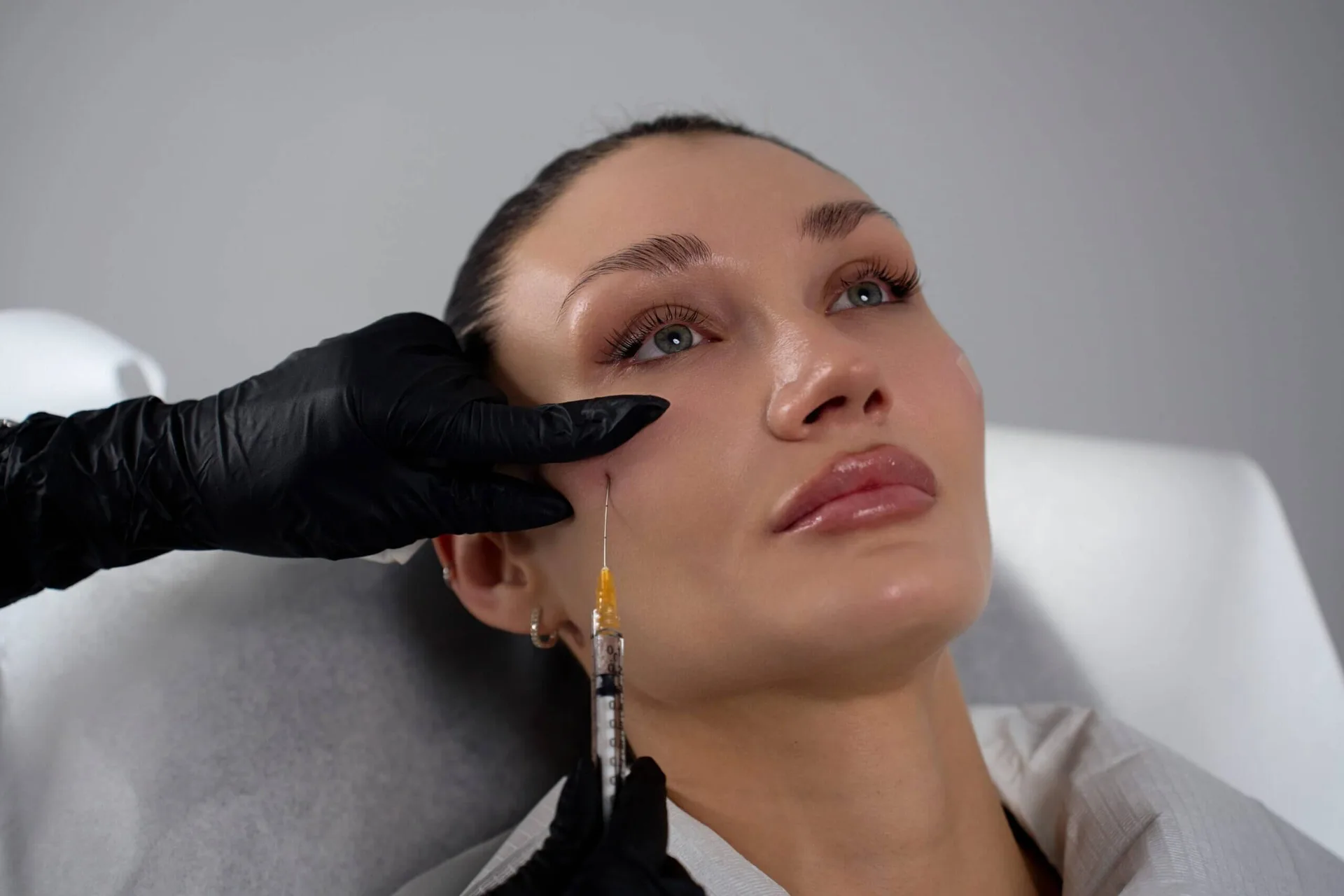 Gloved injector giving a fine-needle injection near the temple and upper cheek of a reclining client